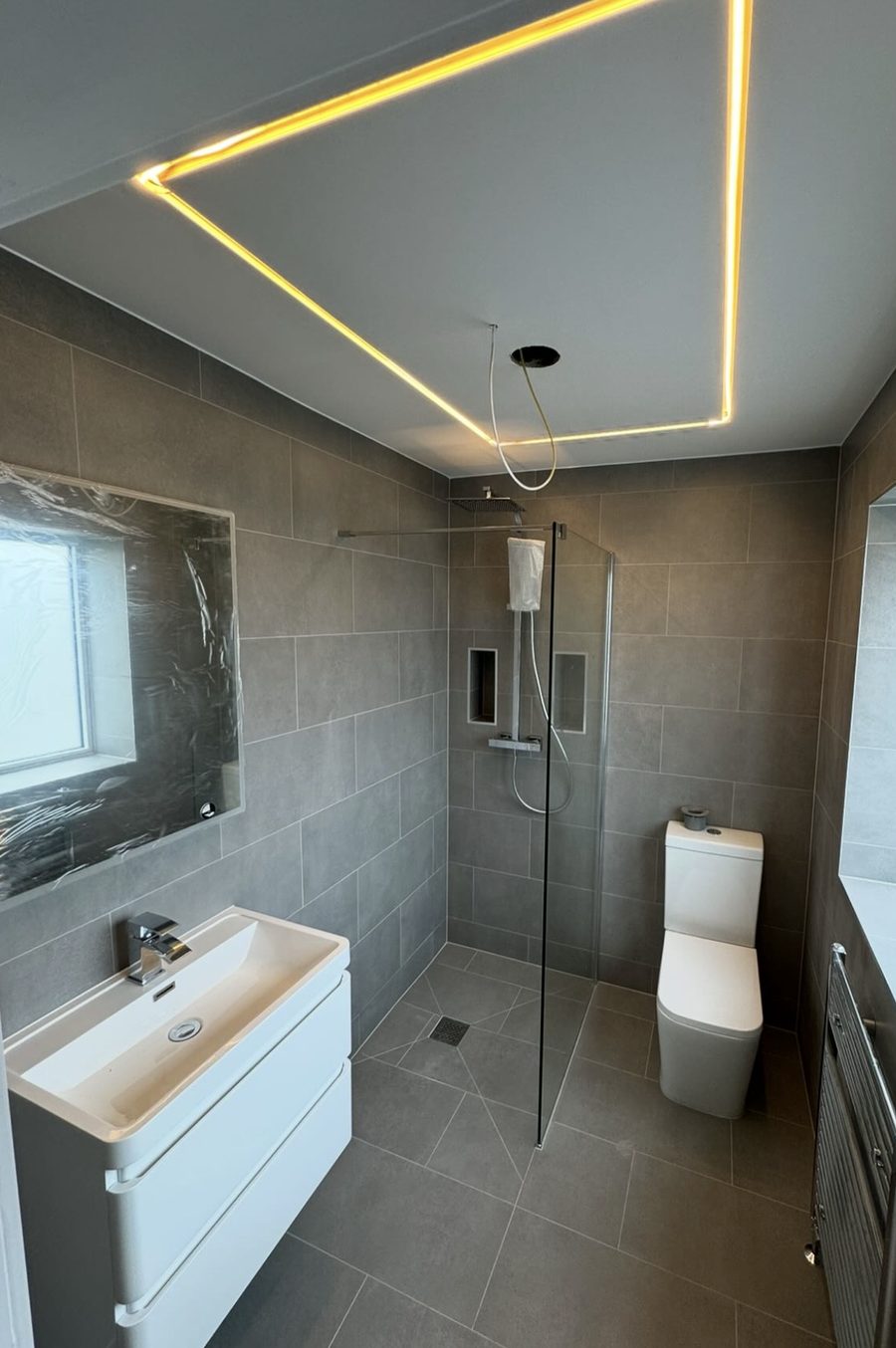 Grey-tiled bathroom with a walk-in shower, LED strip lighting in the ceiling and a wall-hung vanity